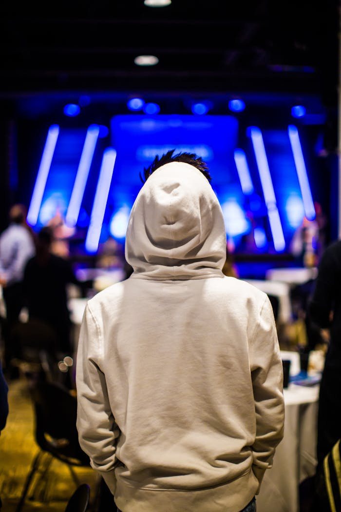 Individual wearing a hoodie attending an indoor event, highlighted by modern blue lighting and blurred crowd.